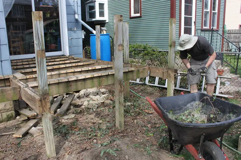 A worker tearing down an old wooden backyard deck, with loose boards and a wheelbarrow of debris