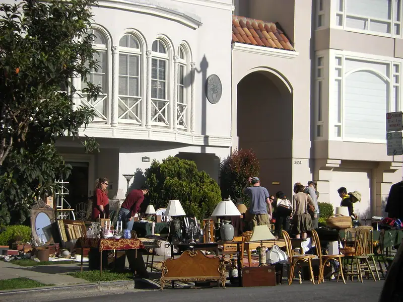 Household furniture, lamps and belongings laid out on a sidewalk in front of a house during a garage sale