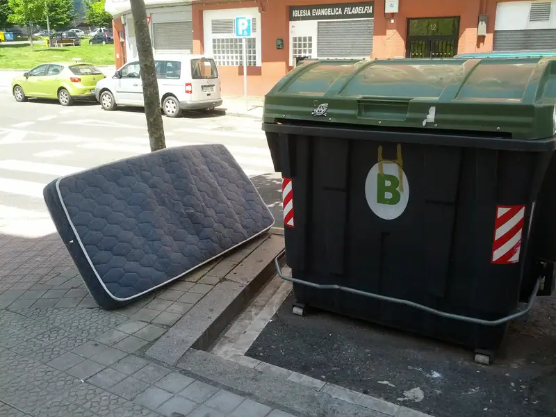 A discarded mattress leaning against a large wheeled trash bin on a city sidewalk