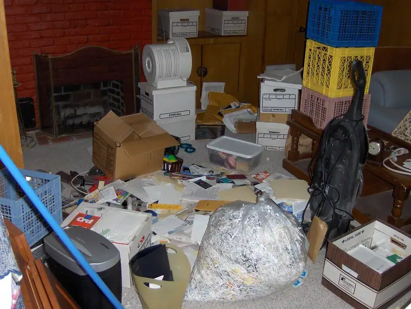 A cluttered room filled with stacked boxes, crates, papers and bags of shredded paper