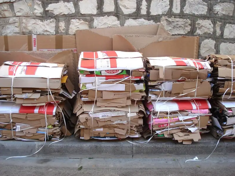 Bundles of flattened cardboard tied with string and stacked against a stone wall for recycling